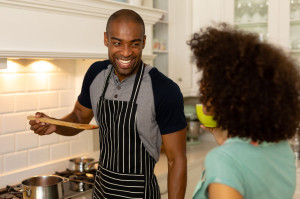 Front view of a mixed race couple standing in the kitchen, the man smiling and offering the woman a taste of food from a wooden spoon
