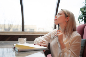 Dreamy young woman writing down future plans and goals in own diary resting in cozy cafeteria