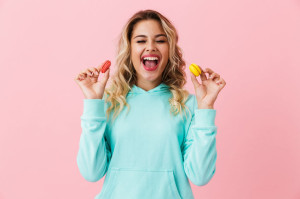 Photo of beautiful woman in basic clothing holding two macaron biscuits, isolated over pink background